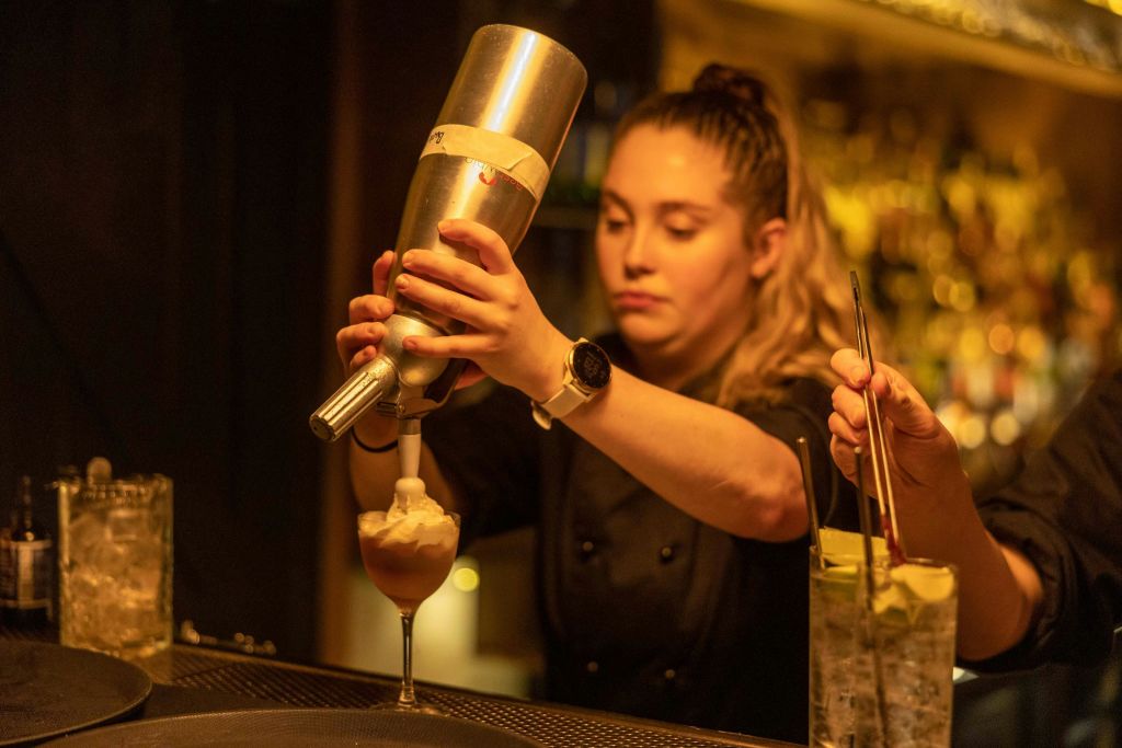 칵테일을 준비하는 바텐더가 사이펀에서 유리잔으로 거품을 내고 근처에 또 다른 음료가 장식되어 있습니다.
