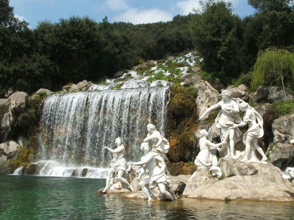 울창한 녹음으로 둘러싸인 계단식 폭포 근처에 사람의 모습이 그려진 하얀 조각상이 서 있습니다.