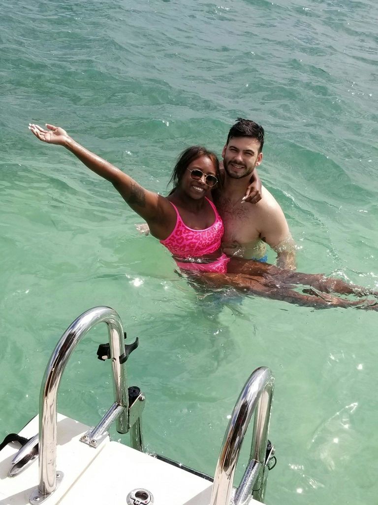 보트 사다리 근처의 맑은 물에 서서 웃고 있는 두 사람, 한 사람은 분홍색 수영복과 선글라스를 착용하고 있습니다.