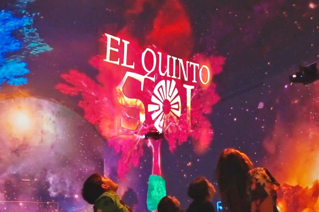 'El Quinto Sol' 이라는 텍스트와 다채로운 시각 효과를 배경으로 생동감 넘치는 디스플레이를 감상하는 사람들.