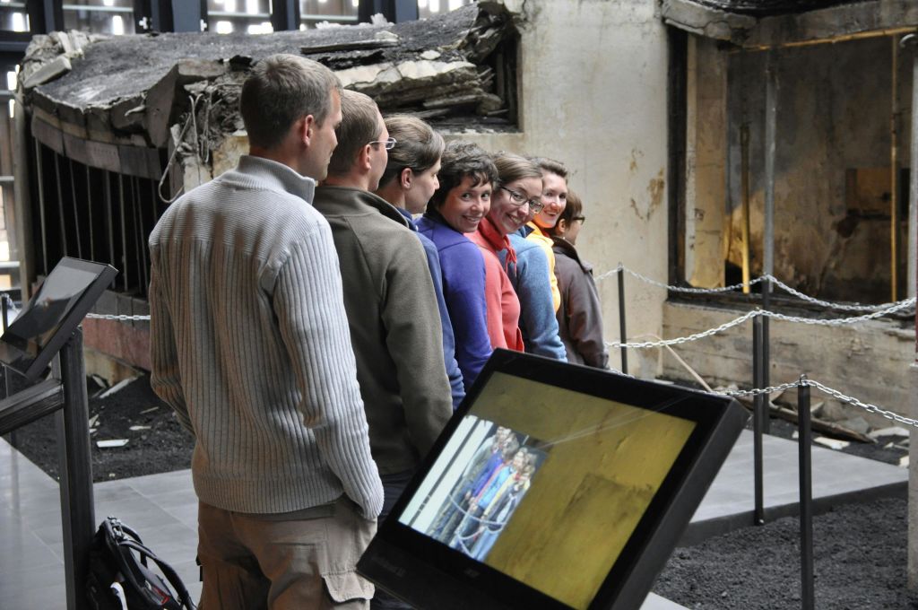 한 무리의 사람들이 실내에 줄을 서서 자신의 이미지가 표시된 화면을 마주보고 있습니다. 잔해와 손상된 벽이 배경에 보입니다.