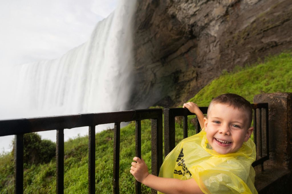 노란 비 판초를 입은 미소 짓는 어린이가 폭포와 바위 절벽을 배경으로 난간 근처에 서 있습니다.