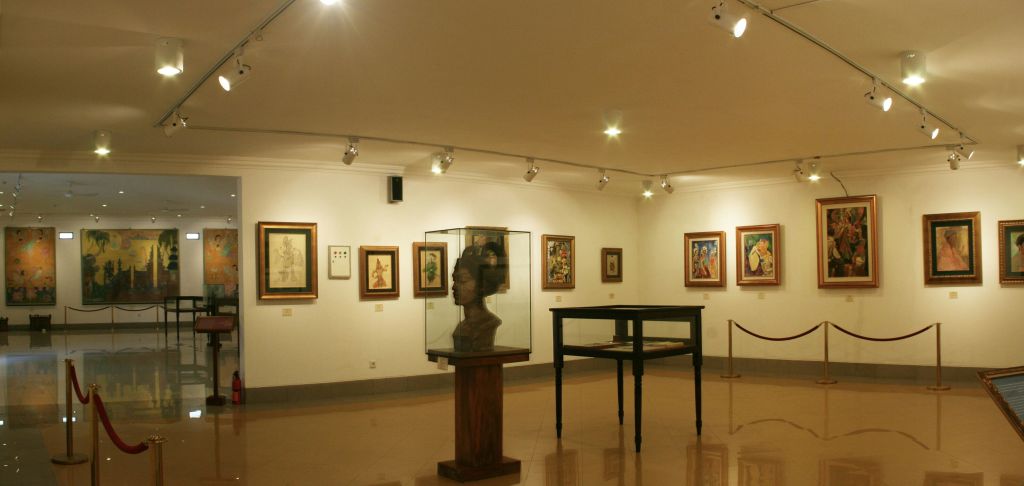 벽에 액자 그림, 나무 받침대 유리 케이스의 흉상, 빈 디스플레이 테이블이 있는 갤러리 내부.