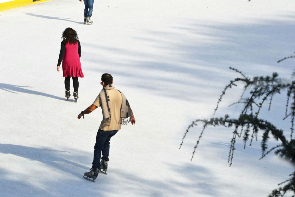 빨간 드레스를 입은 한 여성과 전경에 베이지색 셔츠와 스카프를 두른 남성과 함께 야외 링크에서 스케이트를 타는 사람들.
