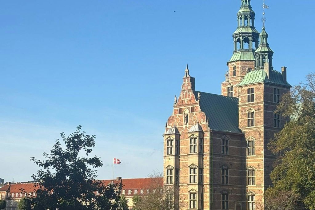 맑고 푸른 하늘 아래 화려한 탑과 붉은 벽돌 외관이 있는 유서 깊은 성을 배경으로 덴마크 국기가 날아옵니다.