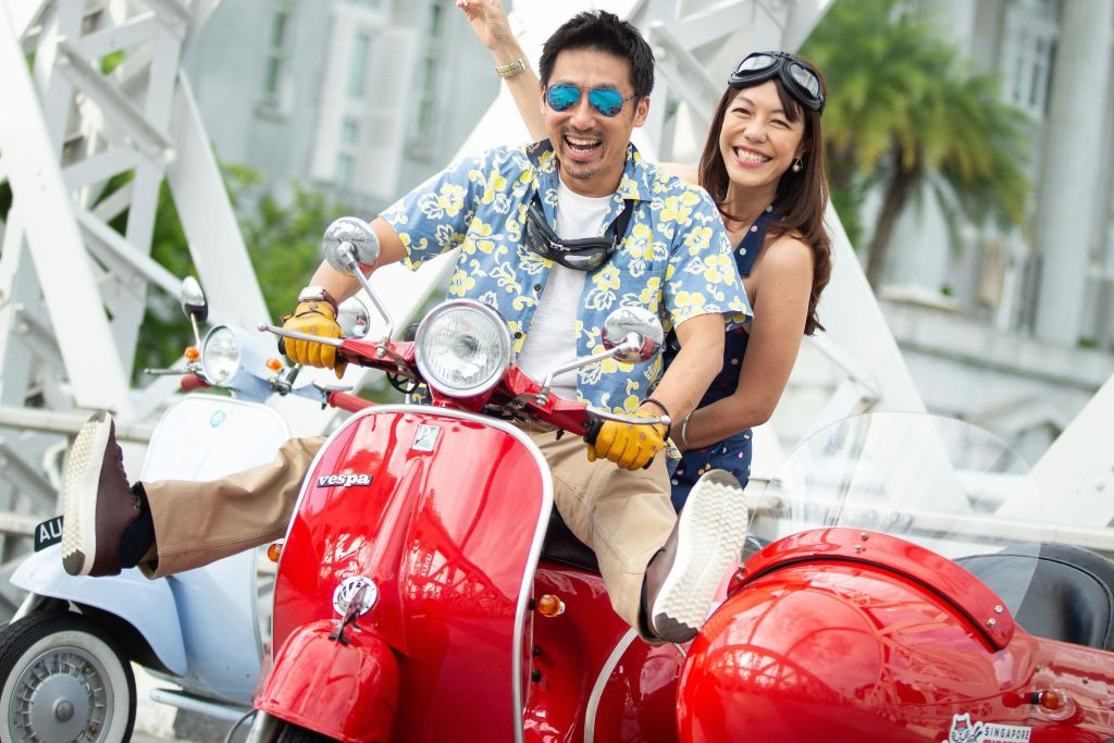 빨간색 스쿠터를 탄 남녀가 하얀 금속 기둥과 건물을 배경으로 평화 표지판을 들고 미소를 짓고 있습니다.