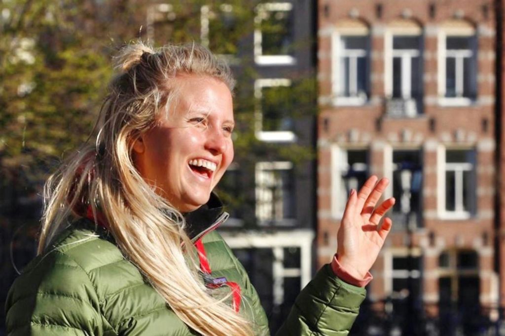 녹색 재킷을 입은 사람이 손동작을 벌리고 활짝 웃고 있습니다. 건물과 나무가 흐릿하게 보입니다.