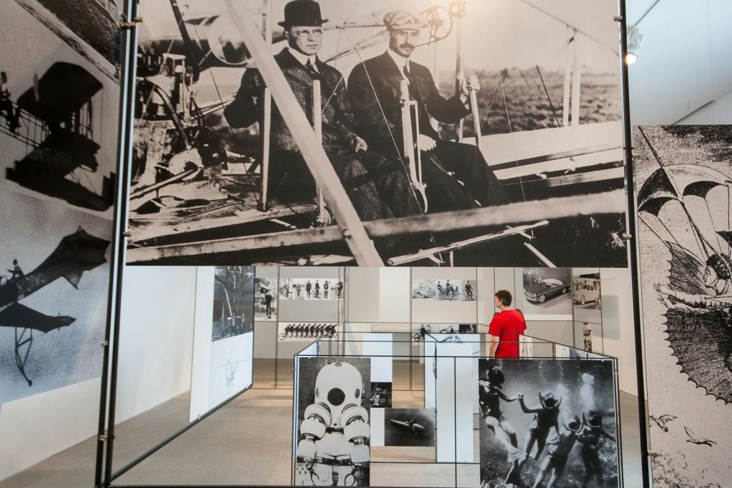 초기 항공 개척자들의 대형 흑백 사진이 전시된 박물관 전시관으로, 빨간 셔츠를 입은 사람이 전시를 관람하고 있습니다.