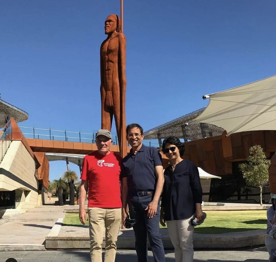 사람의 키가 크고 녹슨 조각상 앞에 세 사람이 서 있습니다. 현대식 건물과 구조물이 배경입니다.