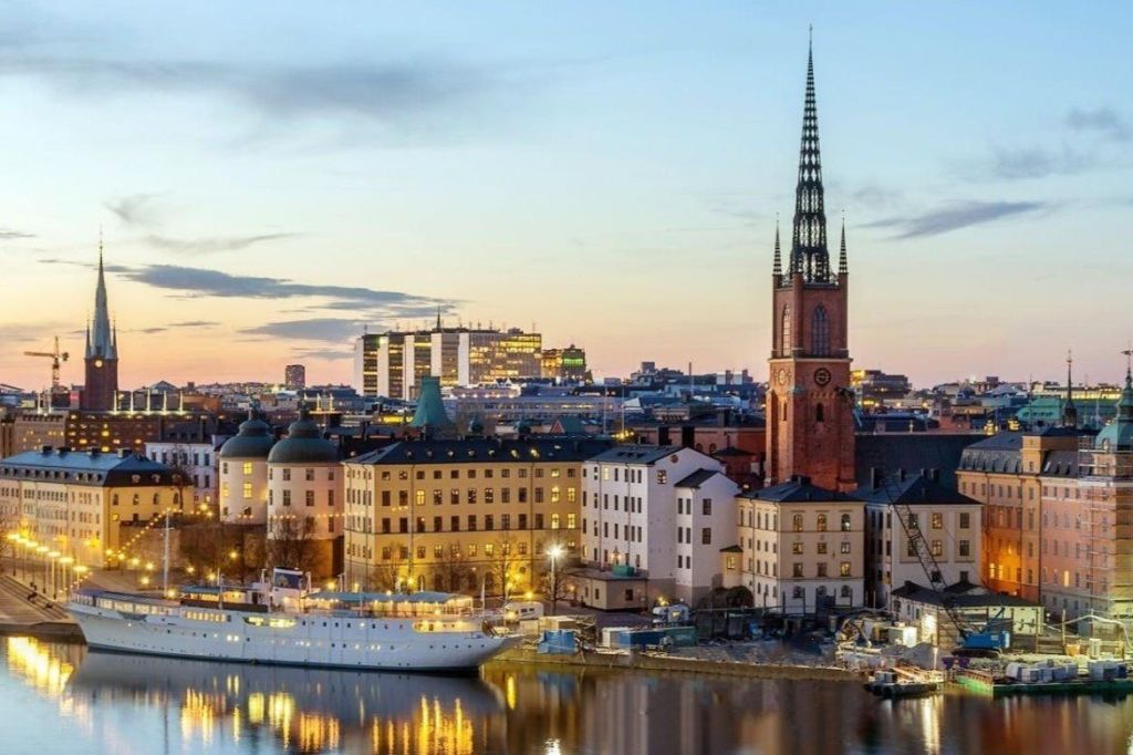 해안가, 역사적인 건물, 높은 첨탑이 있는 교회, 정박된 보트가 있는 석양의 도시 풍경.