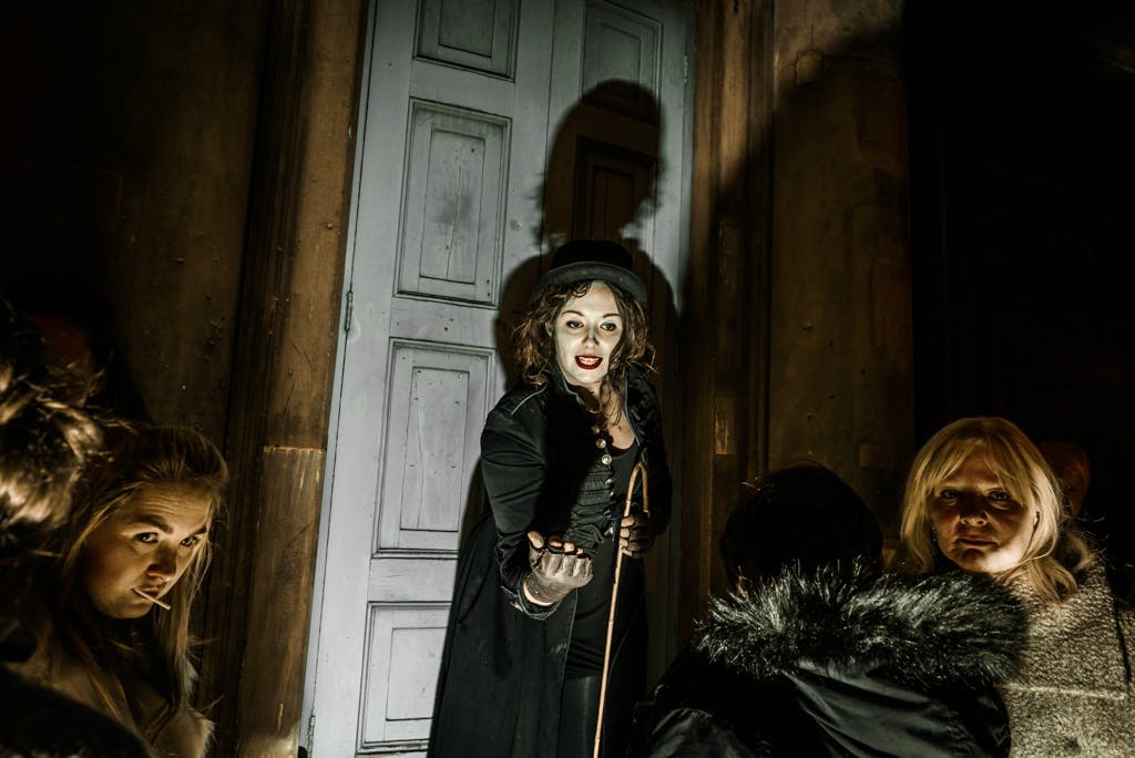 고딕 복장을 한 여성과 상의를 입은 여성이 문 앞에 서서 카메라를 향해 손을 내밀고 있습니다. 어떤 사람들은 그녀의 주위에 보인다.
