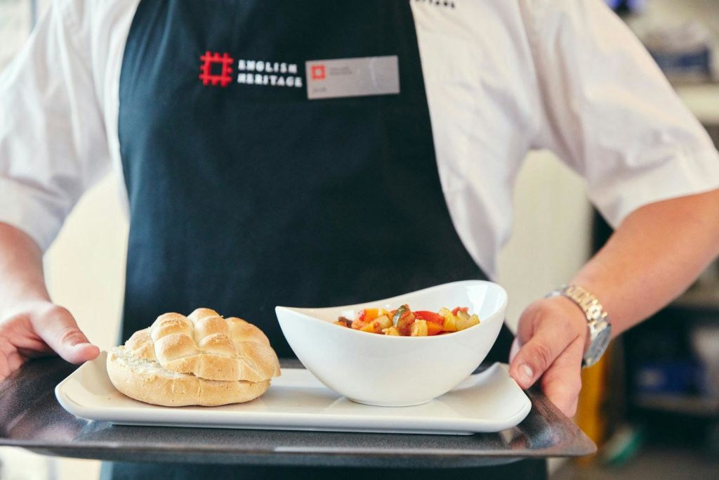 흰색 셔츠와 검은색 앞치마를 입은 웨이터가 야채 스튜 한 그릇과 빵 한 롤이 담긴 트레이를 제공합니다.