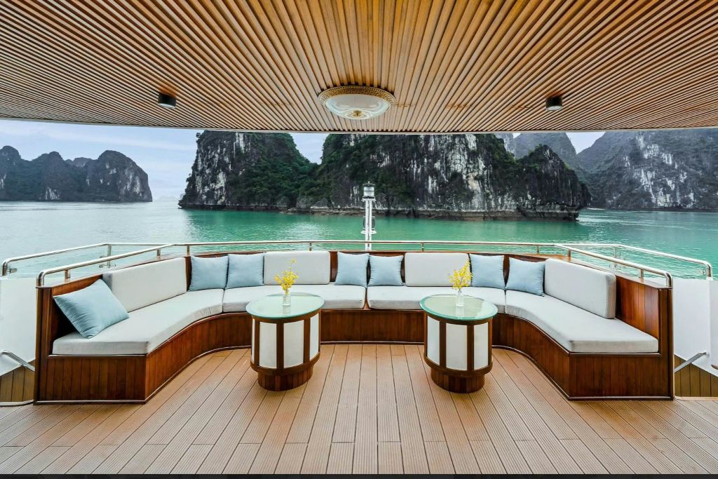 쿠션이 있는 보트 갑판, 꽃이 놓인 원형 테이블 2개, 저 멀리 푸른 물과 바위섬이 내려다보입니다.