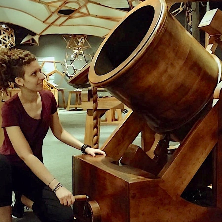 빨간 셔츠를 입은 한 여성이 기하학적 구조물을 배경으로 한 박물관과 같은 환경에서 커다란 나무 망원경과 상호 작용하고 있습니다.