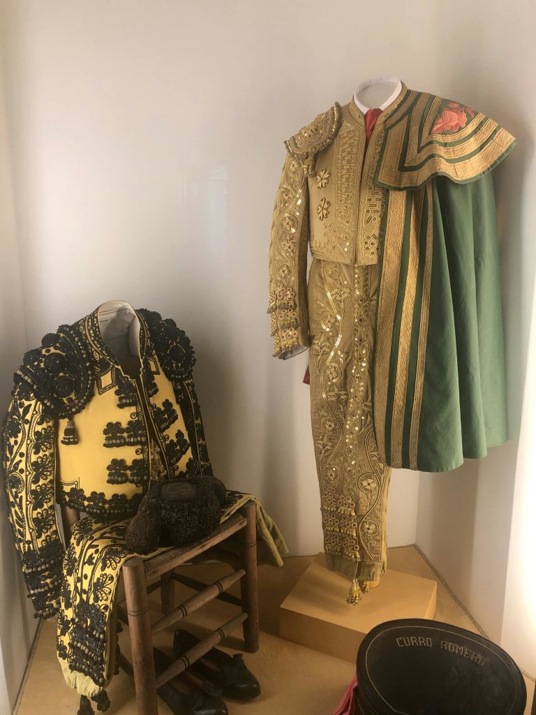 박물관에 전시된 화려한 마타도르 코스튬 두 개. 한 개는 초록색 망토를 두른 채 서 있고, 다른 한 개는 의자에 앉아 있습니다.