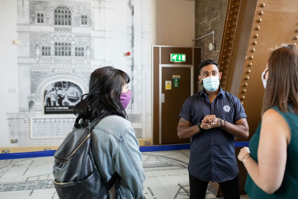 세 사람이 모두 마스크를 착용하고 실내에 서 있습니다. 교복을 입은 남성이 배낭을 메고 있는 여성과 대화하고 있습니다.