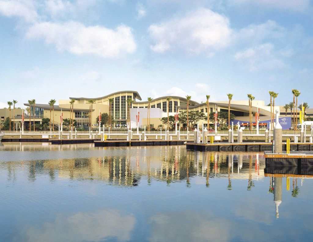 야자수로 둘러싸인 곡선 지붕이 있는 현대적인 건물로, 부분적으로 흐린 하늘 아래 전경에 부두가 있는 잔잔한 물에 반사됩니다.