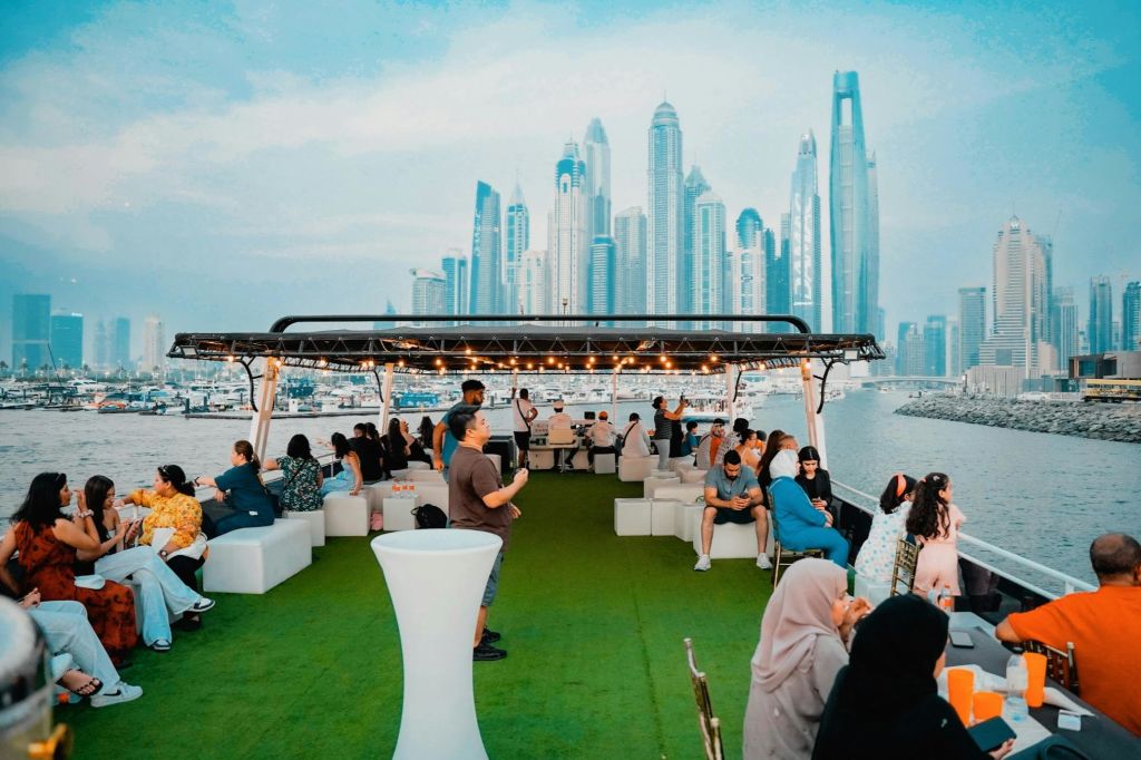 사람들은 인조 잔디 바닥이 깔린 보트에서 높은 건물의 도시 풍경을 배경으로 하얀 소파와 의자에 앉아 있습니다.