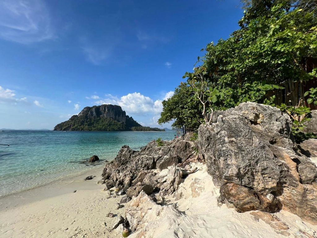 바위 같은 해안선, 맑은 청록색 물, 울창한 초목, 밝고 푸른 하늘 아래 멀리 떨어진 섬이 있는 모래 해변.