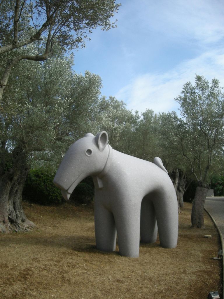 부분적으로 흐린 하늘 아래 마른 풀밭에 올리브 나무로 둘러싸인 양식화 된 동물의 돌 조각.