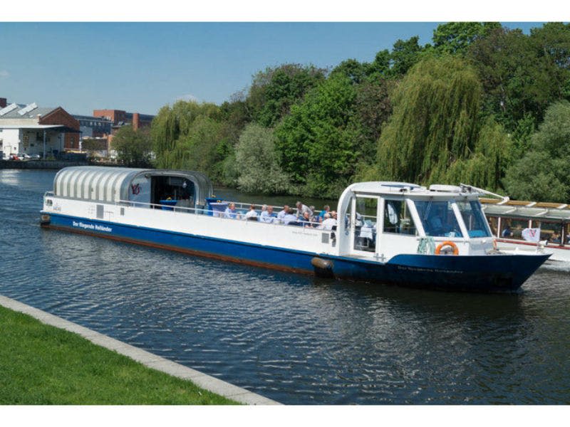 화창한 날에 나무가 늘어선 수로를 따라 승객과 함께 흰색과 파란색 운하 보트가 이동합니다.