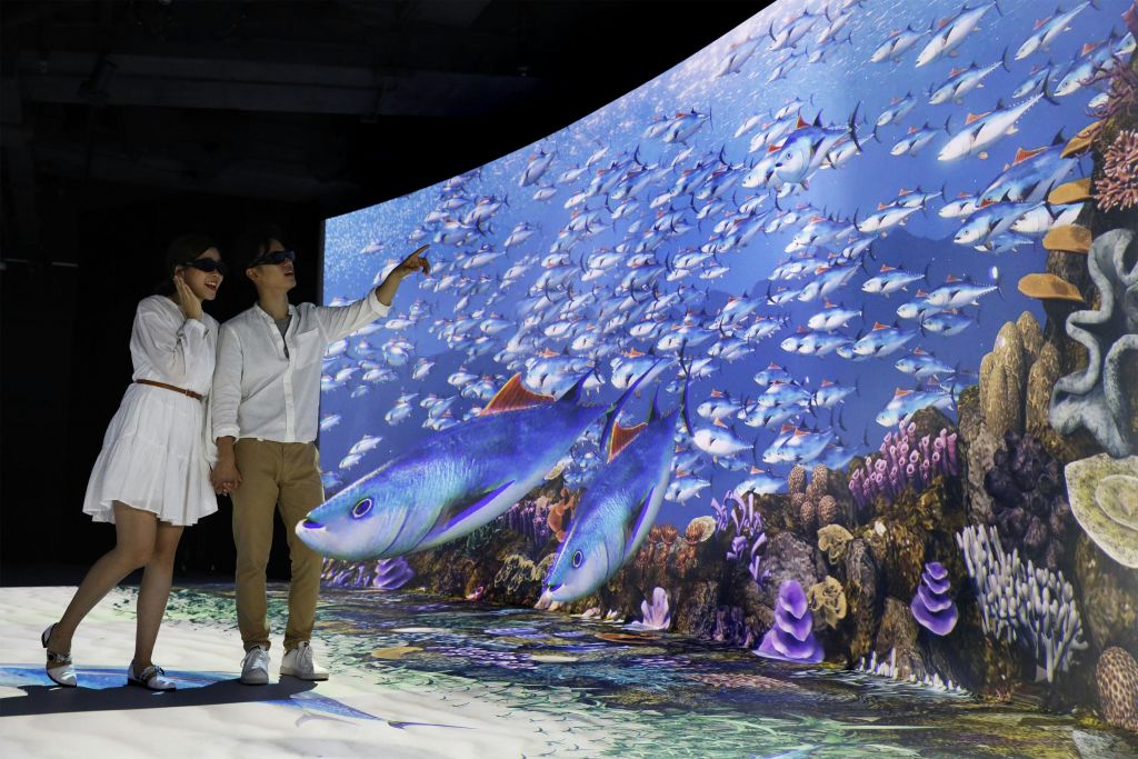 3D 안경을 쓴 두 사람이 물고기와 산호가 있는 수중 장면의 대형 몰입형 디스플레이에 감탄합니다.