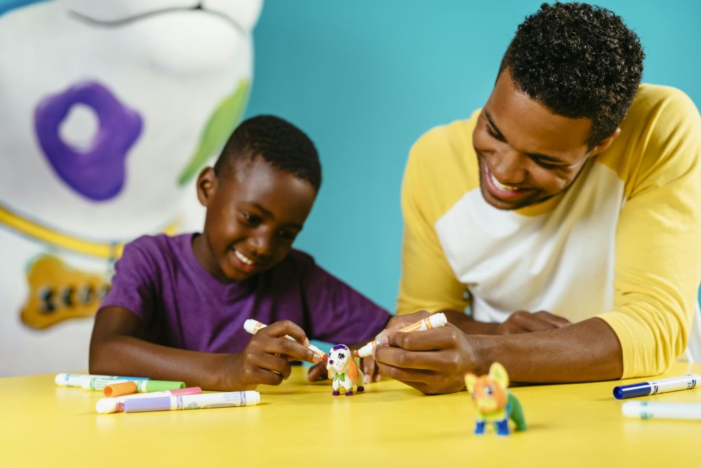 파란색 배경에 노란색 테이블에서 마커가 있는 작은 장난감 피규어를 미소 짓고 색칠하는 두 사람.