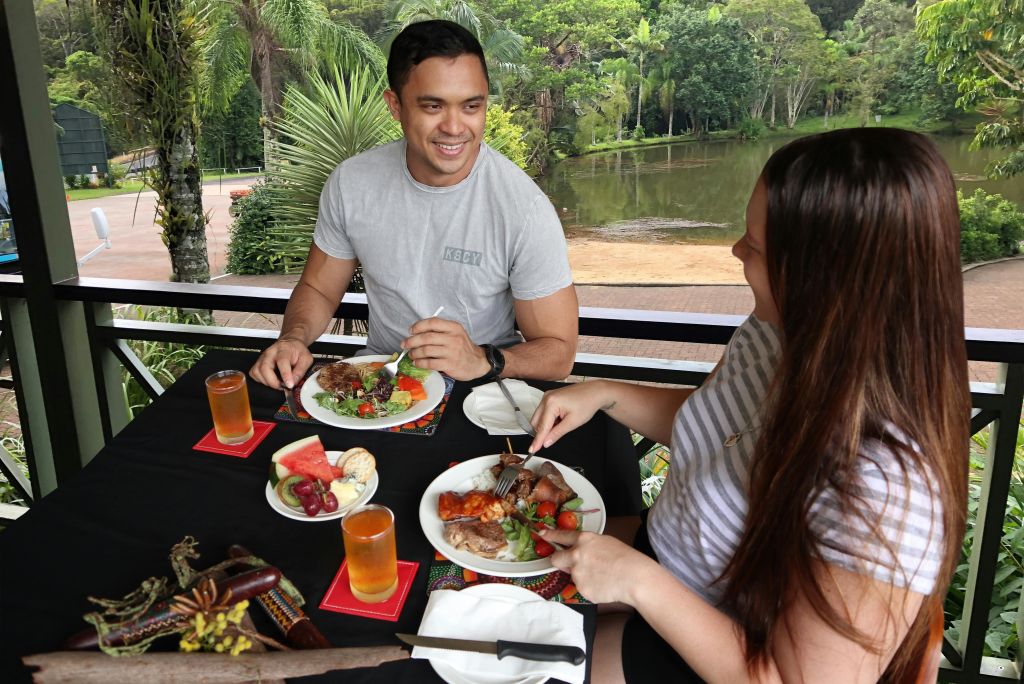 두 사람이 야외 테이블에서 음식, 음료, 과일을 즐기며 식사를 합니다. 울창한 녹음과 연못이 배경으로 보입니다.