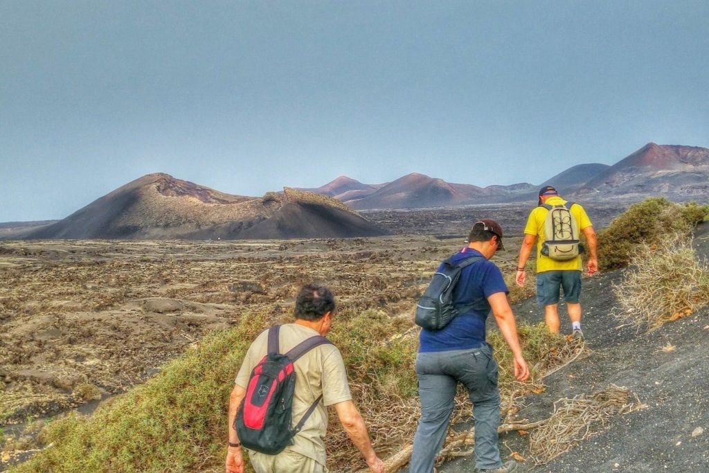 화산 언덕과 맑은 하늘을 배경으로 바위 지형을 걷는 세 명의 등산객.