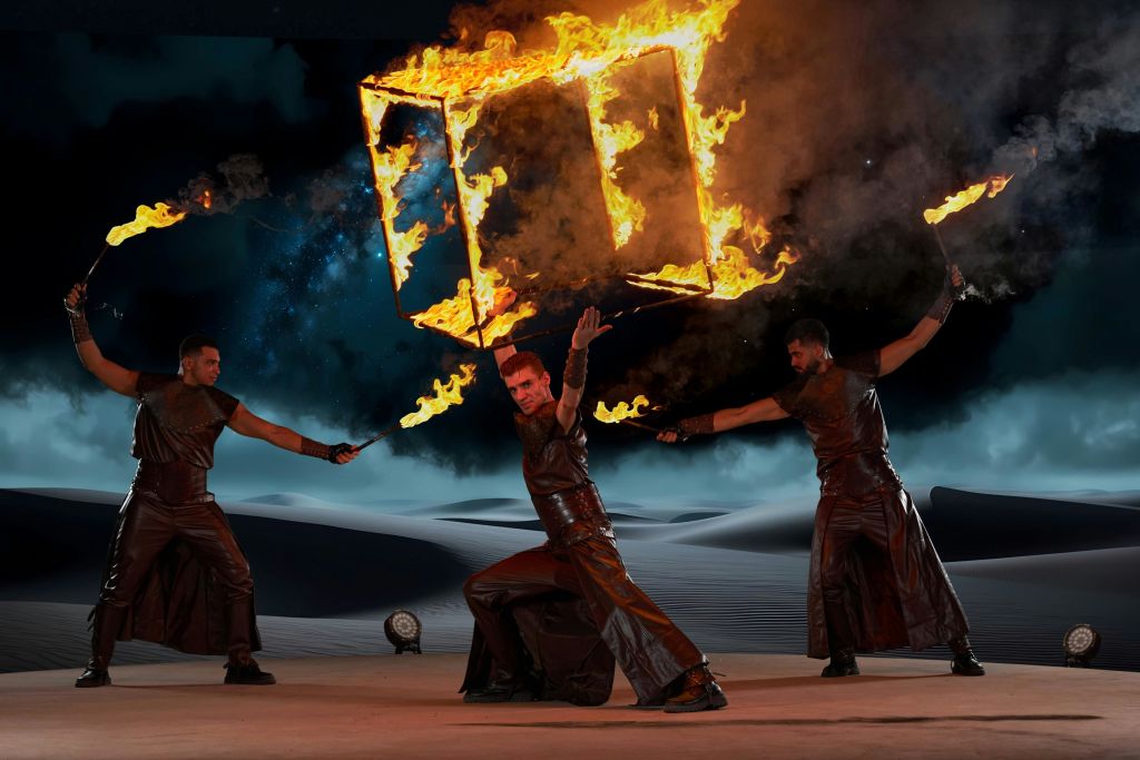 가죽 의상을 입은 두 명의 연주자가 모래 언덕이 있는 어둡고 별이 빛나는 배경에서 불타는 물체를 조작합니다.