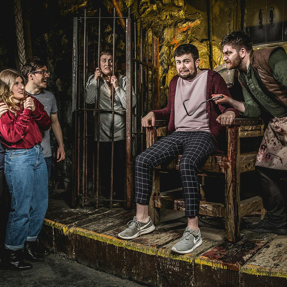 피 묻은 앞치마를 입은 남성이 전기 의자에 앉은 긴장한 남성 근처에서 주사기를 들고 있고, 다른 사람들은 지켜보고 있으며 한 사람이 우리에 갇혀 있습니다.