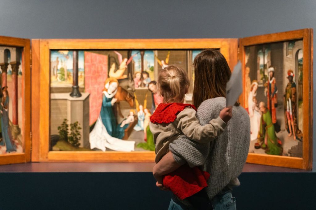 박물관에서 어른과 어린이가 활기찬 트리프티크 그림을 감상합니다. 아이는 흰 종이를 들고 있습니다.