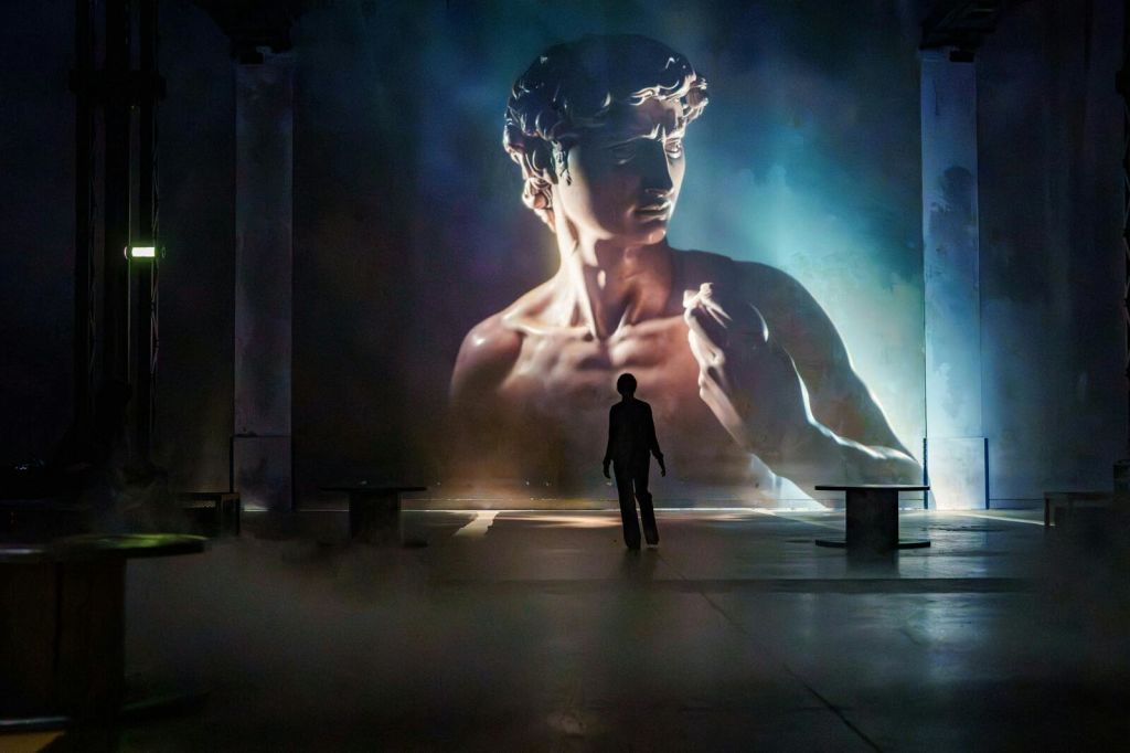 한 사람이 조명이 어두운 방에 서서 극적인 분위기에서 미켈란젤로의 다비드의 커다란 조명 이미지를 마주하고 있습니다.