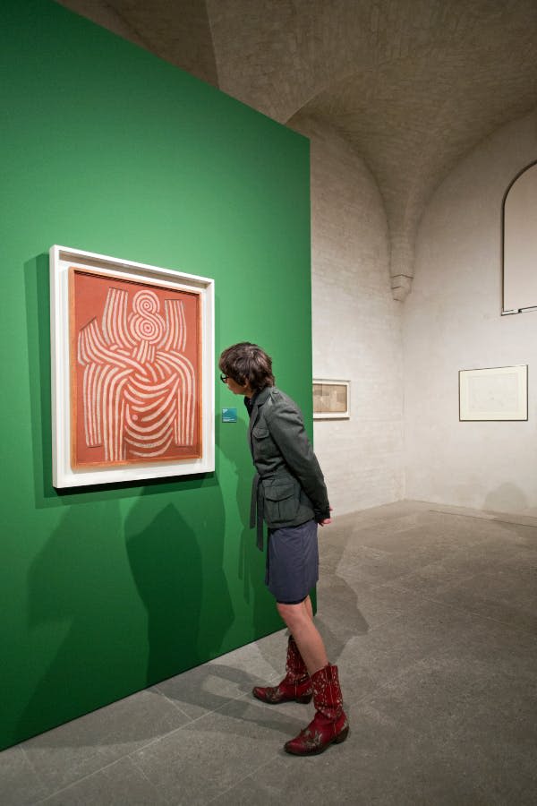 높은 천장과 다양한 액자의 예술품이 있는 미니멀리즘 갤러리 공간의 초록색 벽면에서 추상적인 예술 작품을 감상하는 사람.