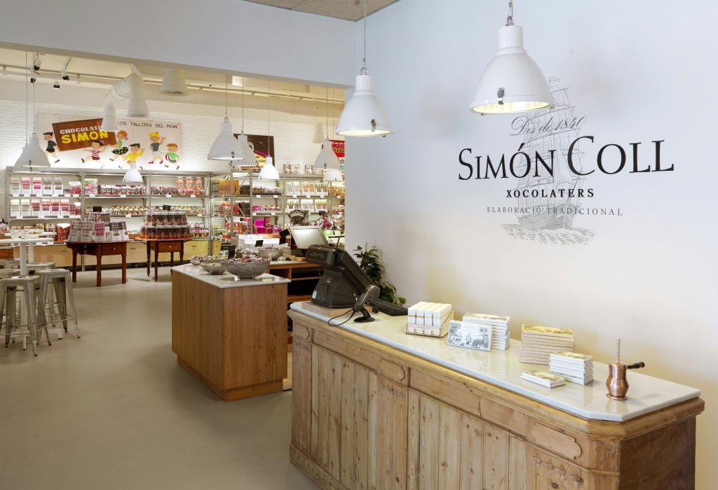 나무 카운터, 금전 등록기, 걸이식 조명, 초콜릿 제품 진열대, "Simón Con." 이라고 적힌 표지판이 있는 상점 내부