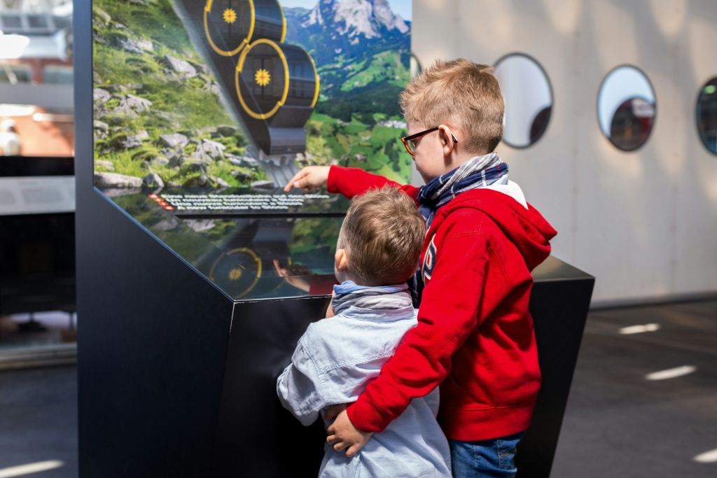 두 소년이 원형 창문이 있는 방 안의 전시물을 바라보며 가리키고 있습니다. 한 소년이 빨간색 옷을 입고 있습니다.