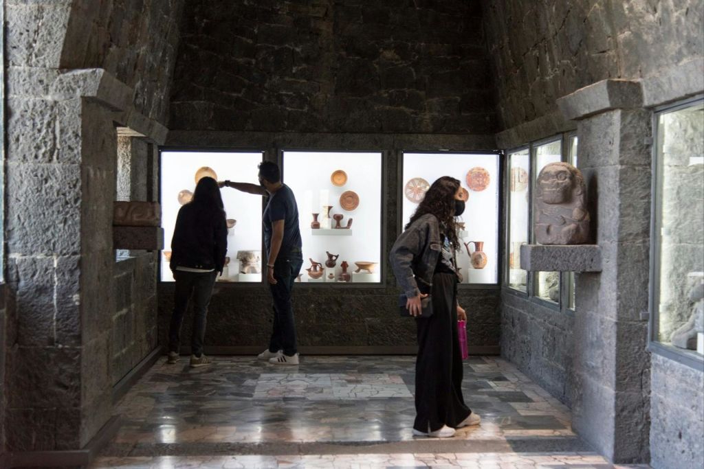 조명이 희미한 돌 벽 박물관 전시관에 전시된 고대 도자기와 유물을 관찰하는 사람들.