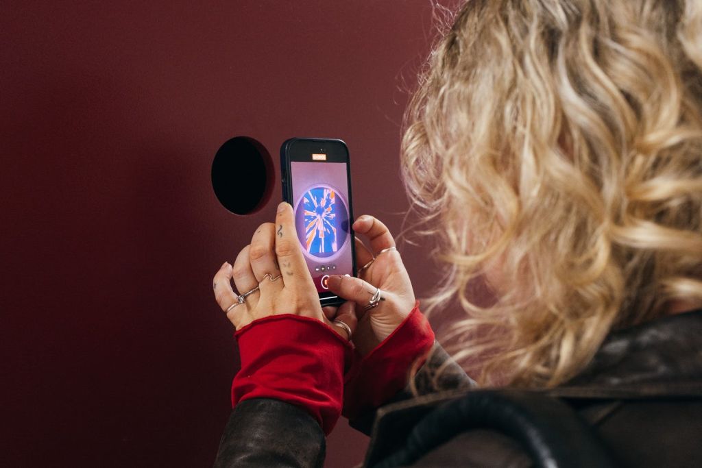 곱슬머리 금발의 사람이 빨간 벽의 원형 구멍을 통해 다채로운 이미지를 찍기 위해 스마트폰을 사용합니다.