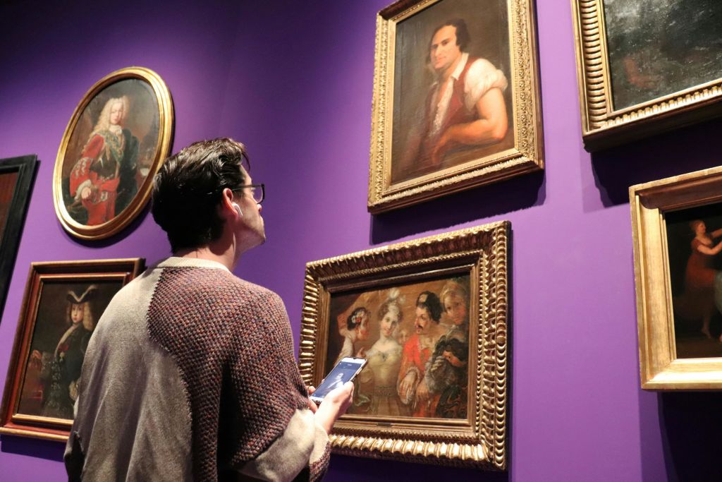 스마트폰을 들고 있는 사람이 갤러리의 자주색 벽에 걸린 액자 그림을 관찰합니다.