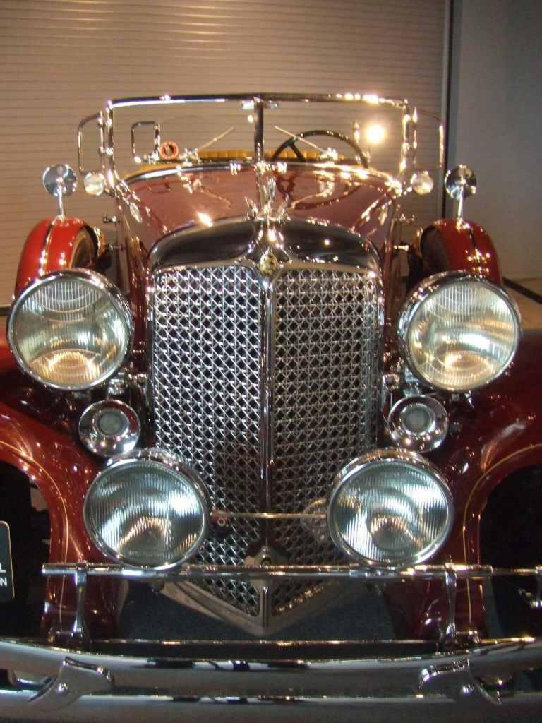 반짝이는 그릴, 4개의 헤드라이트, 반사되는 마룬 바디가 있는 빈티지 자동차의 전면 모습.