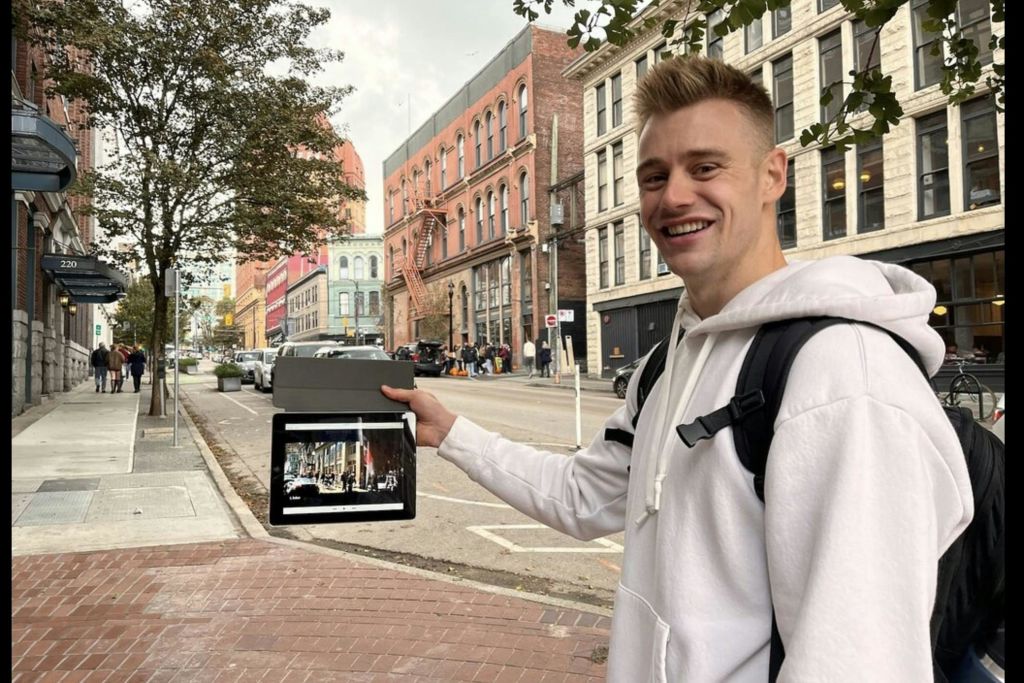 유서 깊은 건물이 늘어선 거리에서 태블릿을 들고 있는 흰색 후드티를 입은 미소 짓는 사람.