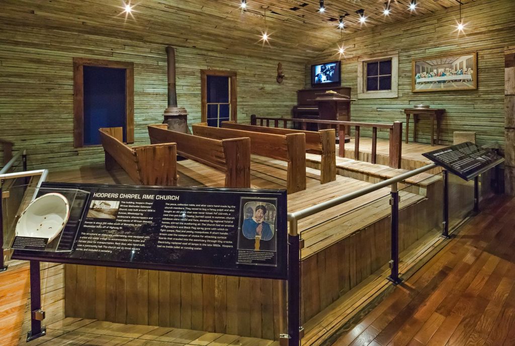 벤치, 화목 난로, 역사적 전시 표지판, TV, 피아노가 있는 목조 예배당 내부. "Hoopers Chapel AME Church" 라는 표지판.
