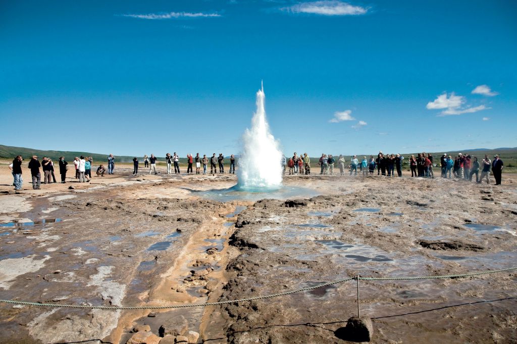 청명한 푸른 하늘 아래에서 분출하는 간헐천을 배경으로 바위 지형과 드문드문 녹음으로 둘러싸인 간헐천을 관찰하는 사람들.