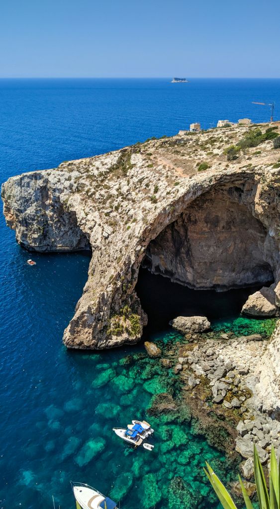 푸른 바다 위에 해안 절벽 아치, 맑은 물과 아래 바위, 바다의 작은 보트, 절벽 위의 먼 건물.