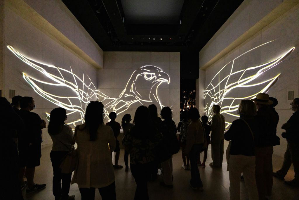 한 무리의 사람들이 새의 날개가 빛나는 윤곽선과 벽에 투영된 새 머리가 있는 희미한 조명으로 꾸며진 방에 서 있습니다.