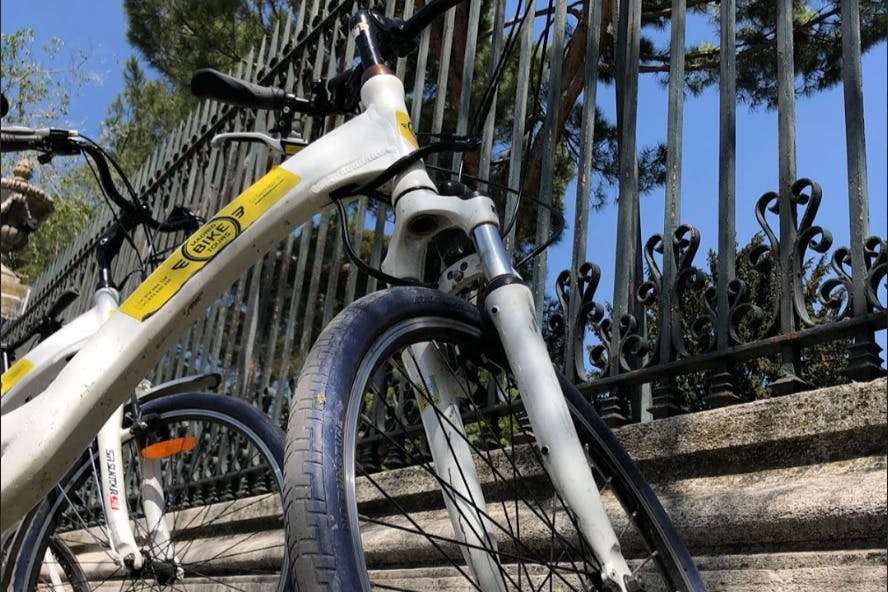 화려한 디테일이 돋보이는 금속 울타리 옆에 노란색 스티커가 부착된 흰색 자전거의 클로즈업. 나무가 배경으로 보입니다.