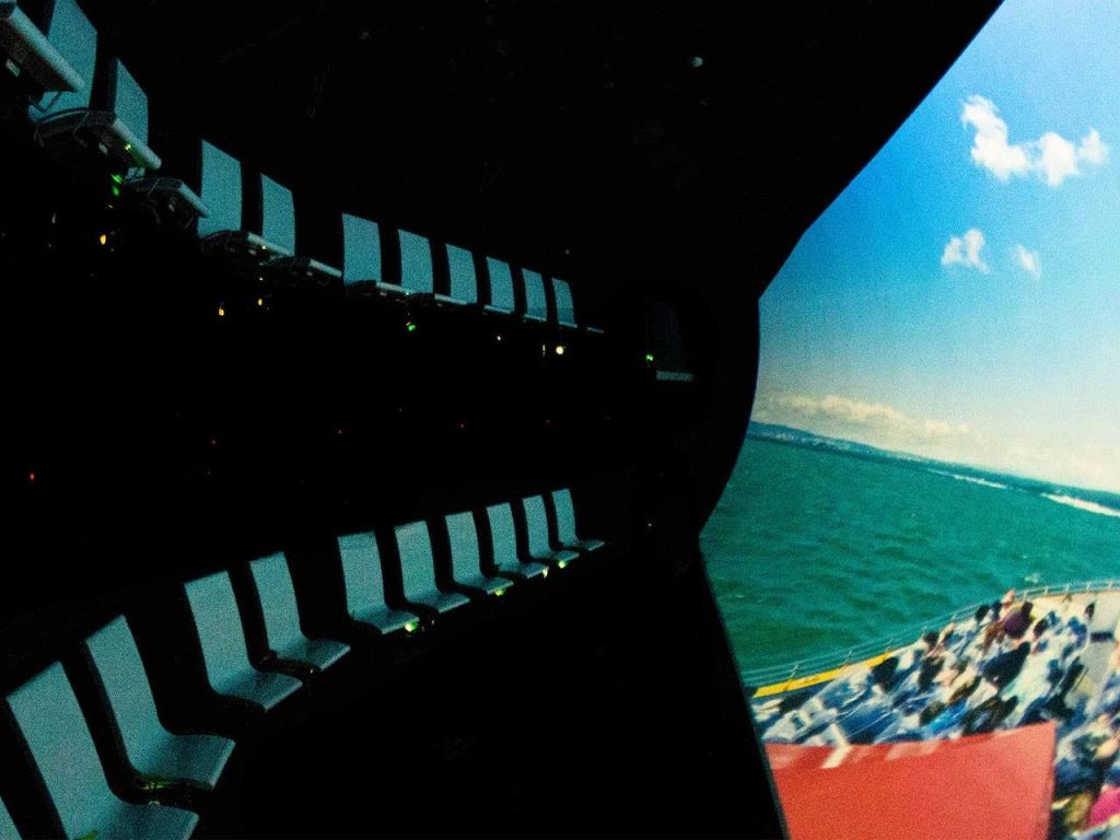 푸른 하늘 아래 바다의 장면을 보여주는 커다랗고 구부러진 화면을 마주하고 있는 빈 좌석이 줄지어 있습니다.