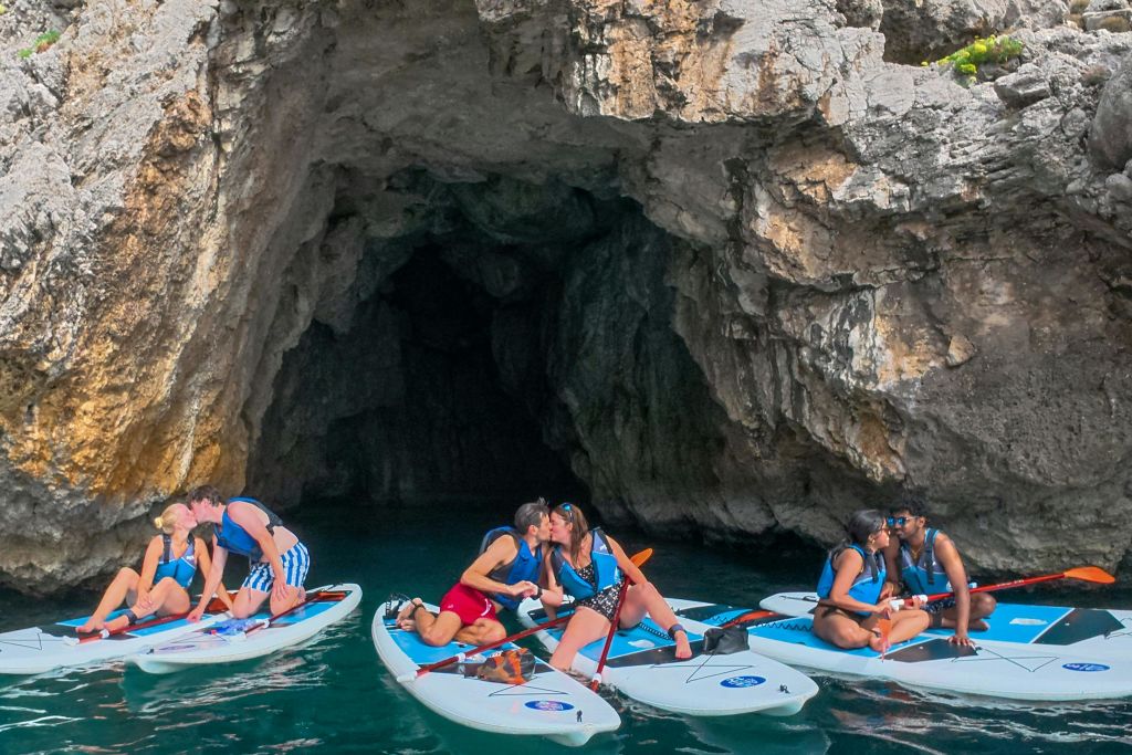 한 무리의 사람들이 바위 동굴 근처에서 패들보드를 타고 있으며, 일부 커플은 키스하고 다른 커플은 활동을 즐깁니다.