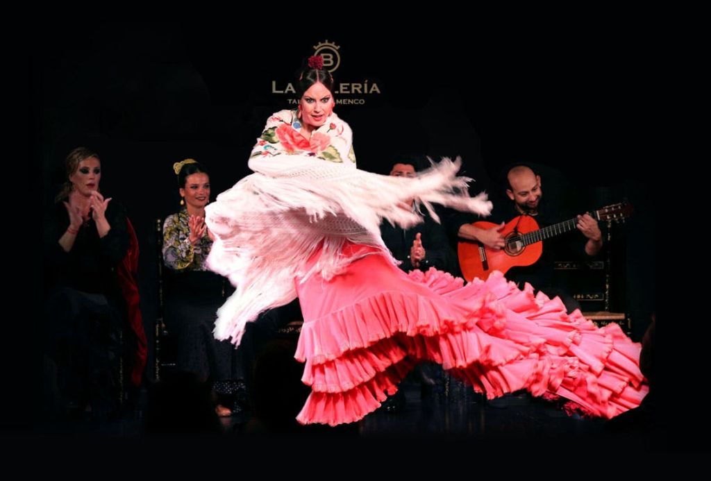 분홍색 드레스와 프린지 숄을 입은 플라멩코 댄서가 무대에서 활기차게 공연하며, 좌석에 앉은 사람들이 배경에서 박수를 치고 있습니다.