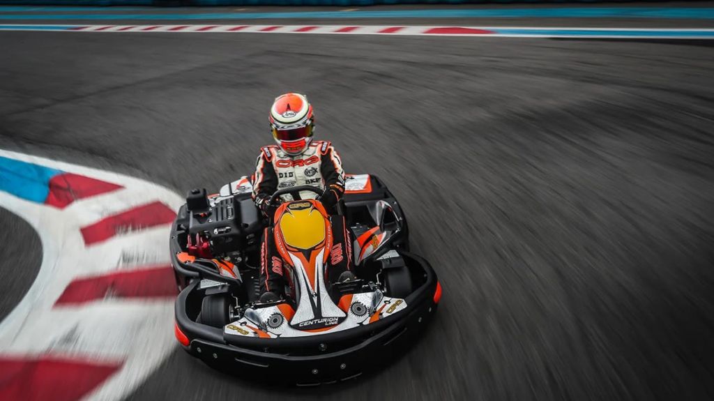 레이싱 장비와 헬멧을 착용한 사람이 트랙 코너에서 고카트를 운전합니다. 고카트에는 화려한 데칼과 브랜딩이 있습니다.